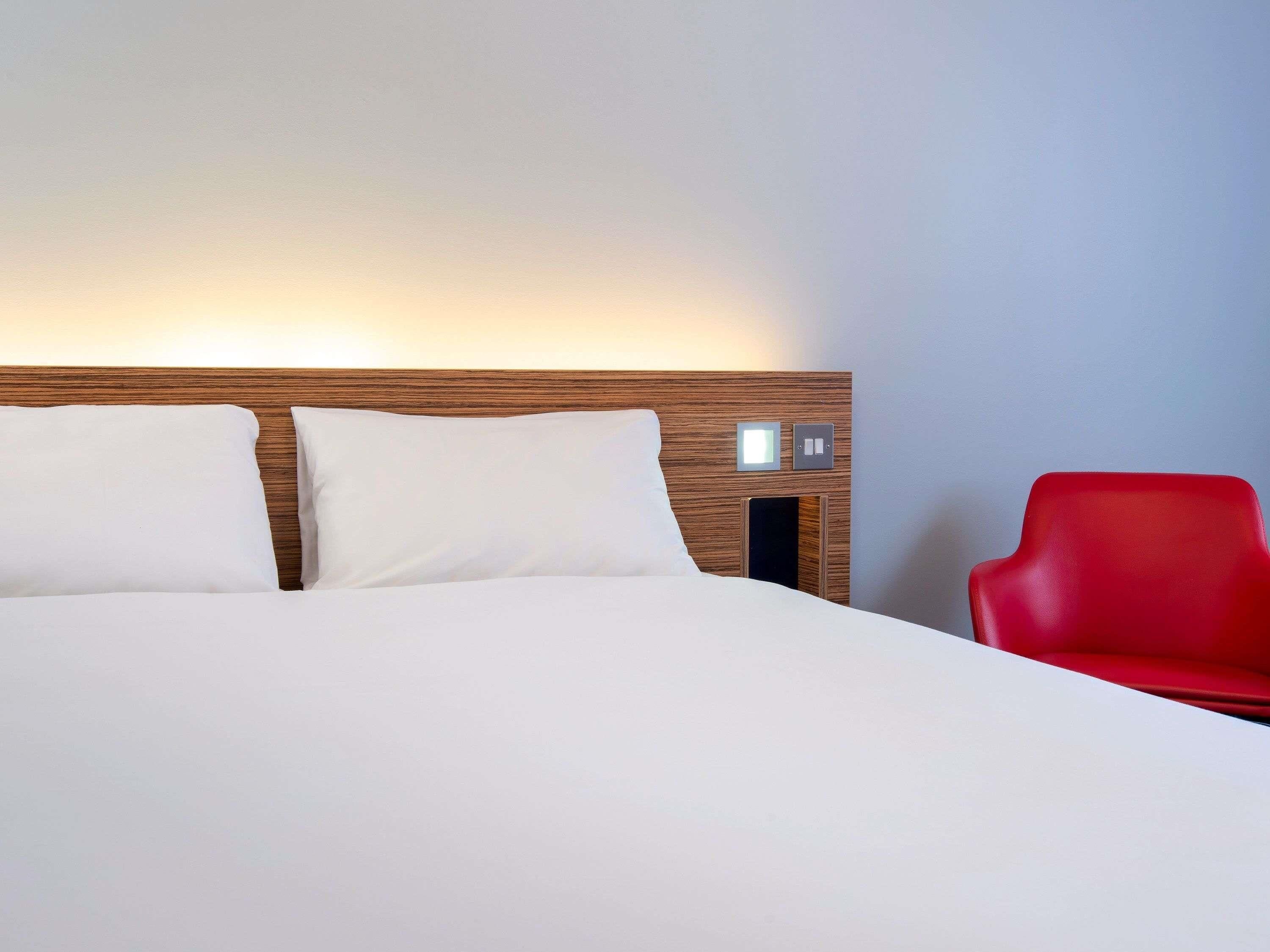 Ein Hotelzimmer mit einem hölzernen Kopfteil, zwei weißen Kissen, einem roten Sessel und einer Wandleuchte.