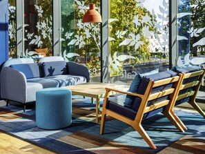 A modern interior with a sofa, ottoman, and wooden chair, large windows with decorative glass, and a hanging pendant light. - Ibis budget London Heathrow Central (Hounslow)