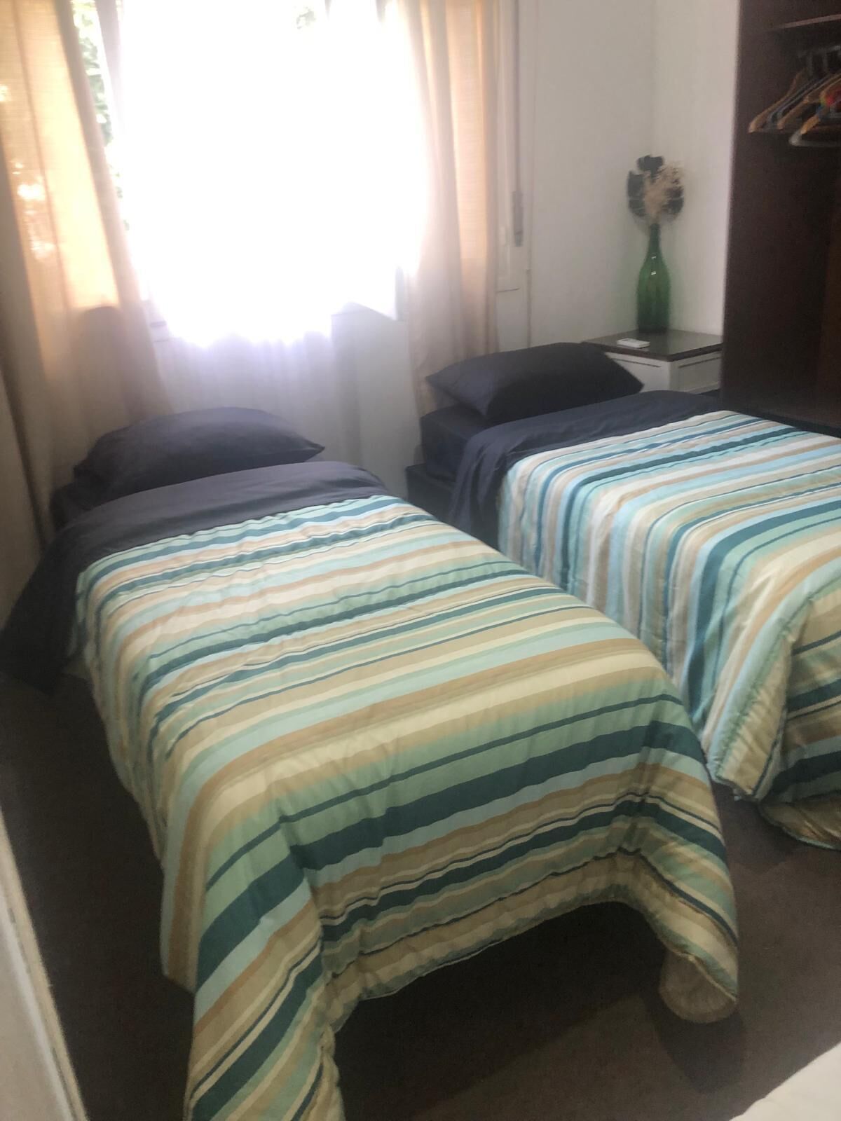 A small, compact room with two single beds, a nightstand, a green vase, and a window with curtains.
