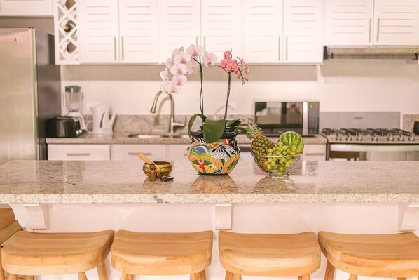 A kitchen with white cabinets, a granite countertop, a blender, a coffee maker, a decorative bowl, and a pineapple.