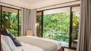 Un dormitorio moderno con una cama grande, una mesita de noche y vistas a exuberante vegetación a través de amplias ventanas.