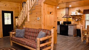 A wooden cabin interior with a plaid sofa, a staircase, a kitchen area, and a dining table.