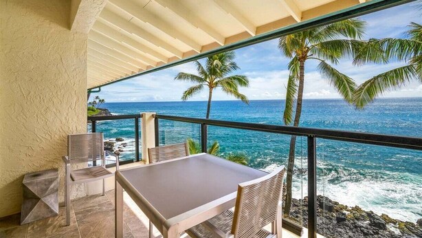 A balcony with a table and chairs, overlooking a coastal view with palm trees and the ocean. - Poipu Shores 402A - Full A/C Luxury, Sublime Views (Koloa)
