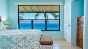 A bedroom with a large window overlooking the ocean, a bed with patterned bedding, a wooden dresser, and a small nightstand with a lamp. - Poipu Shores 402A - Full A/C Luxury, Sublime Views (Koloa)