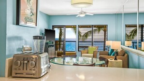 A room with a kitchenette, a glass table, a ceiling fan, and a view of the ocean. - Poipu Shores 402A - Full A/C Luxury, Sublime Views (Koloa)