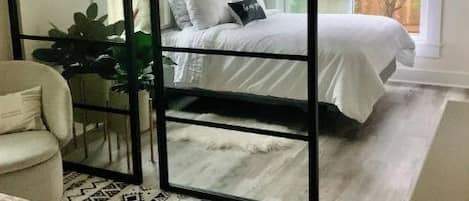 Un dormitorio con una cama grande, un separador con espejo, una silla blanca y una alfombra estampada.