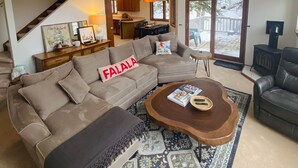 Un salon spacieux avec un plafond haut, un escalier en bois, un grand canapé agrémenté d’un plaid, une table basse et une vue sur l’extérieur grâce à une porte-fenêtre coulissante.