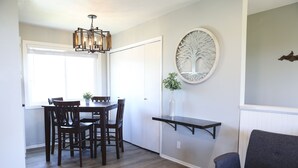 A dining area with a table and chairs, a hanging light fixture, a decorative wall piece, and a small shelf with a plant. - Over the Oaks - Great View - Whole Home - Stylish  (Thousand Oaks)
