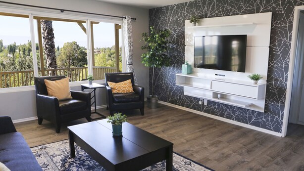 A modern living room with a large TV, dark wood flooring, a patterned rug, and a balcony with a view. - Over the Oaks - Great View - Whole Home - Stylish  (Thousand Oaks)