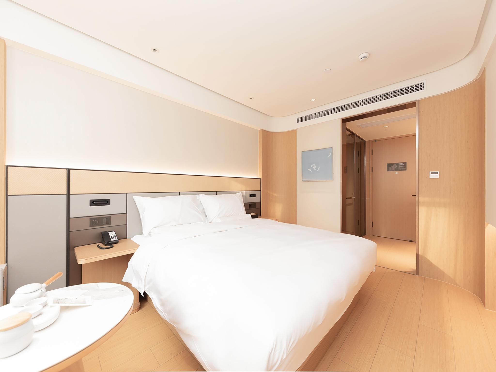 A modern hotel room with a large bed, a bedside table with a phone, a small table with a kettle, and a chair.