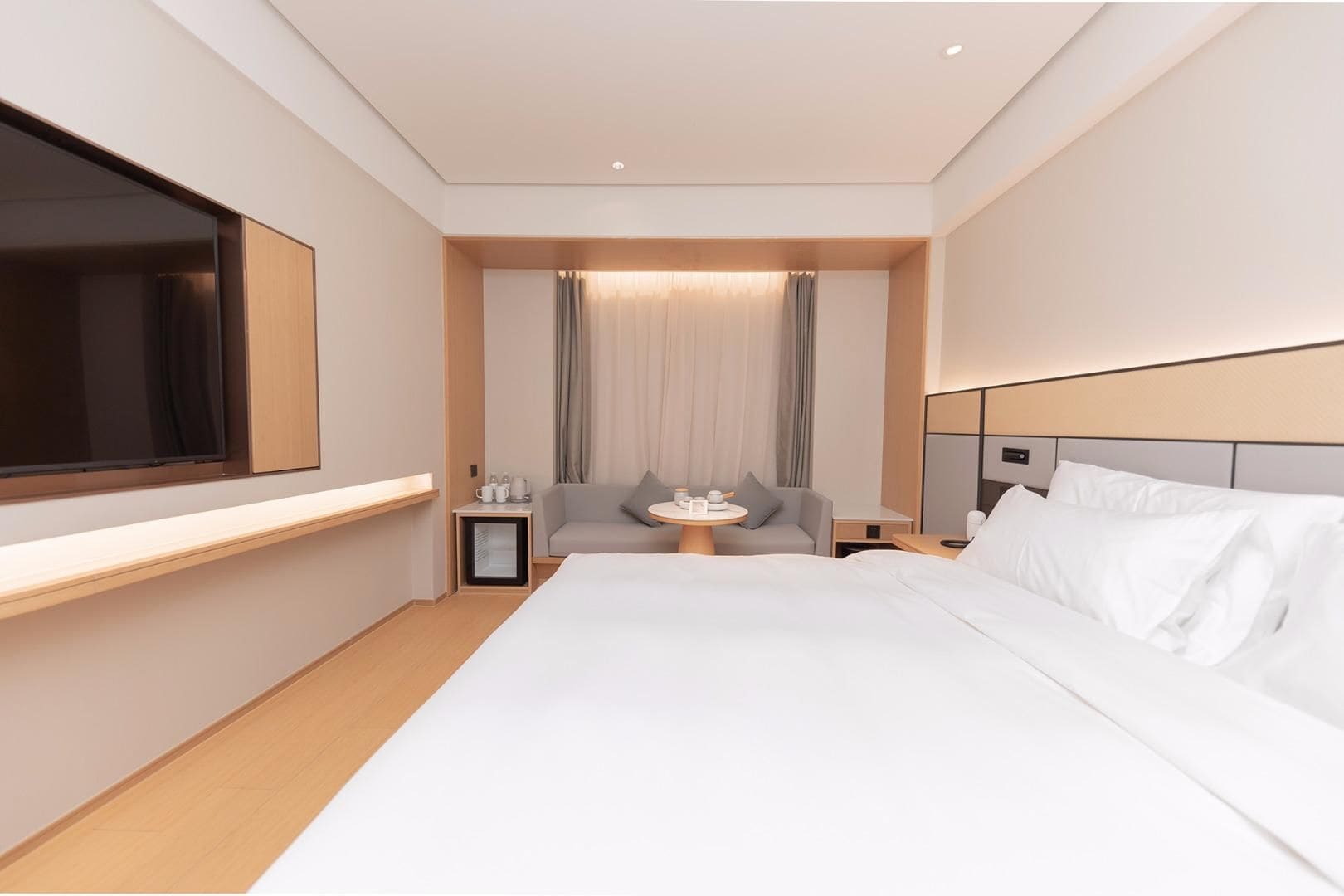 Una habitación de hotel moderna con una cama grande, un televisor de pantalla plana, una mesita de noche con lámpara y una zona de estar con mesa y sillas.