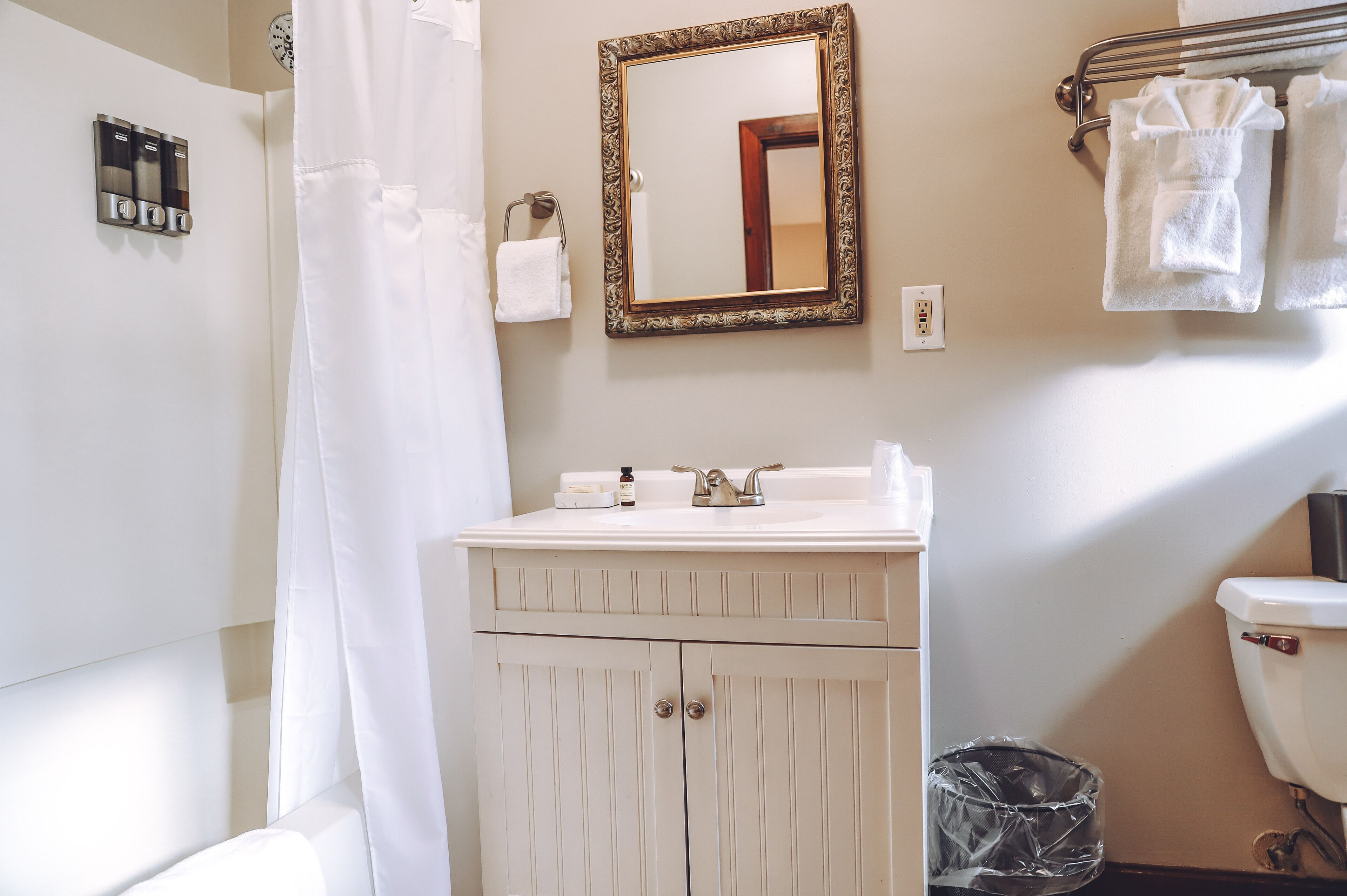 A bathroom with a white sink, a mirror with a decorative frame, a towel rack with towels, a shower curtain, and a trash bin.