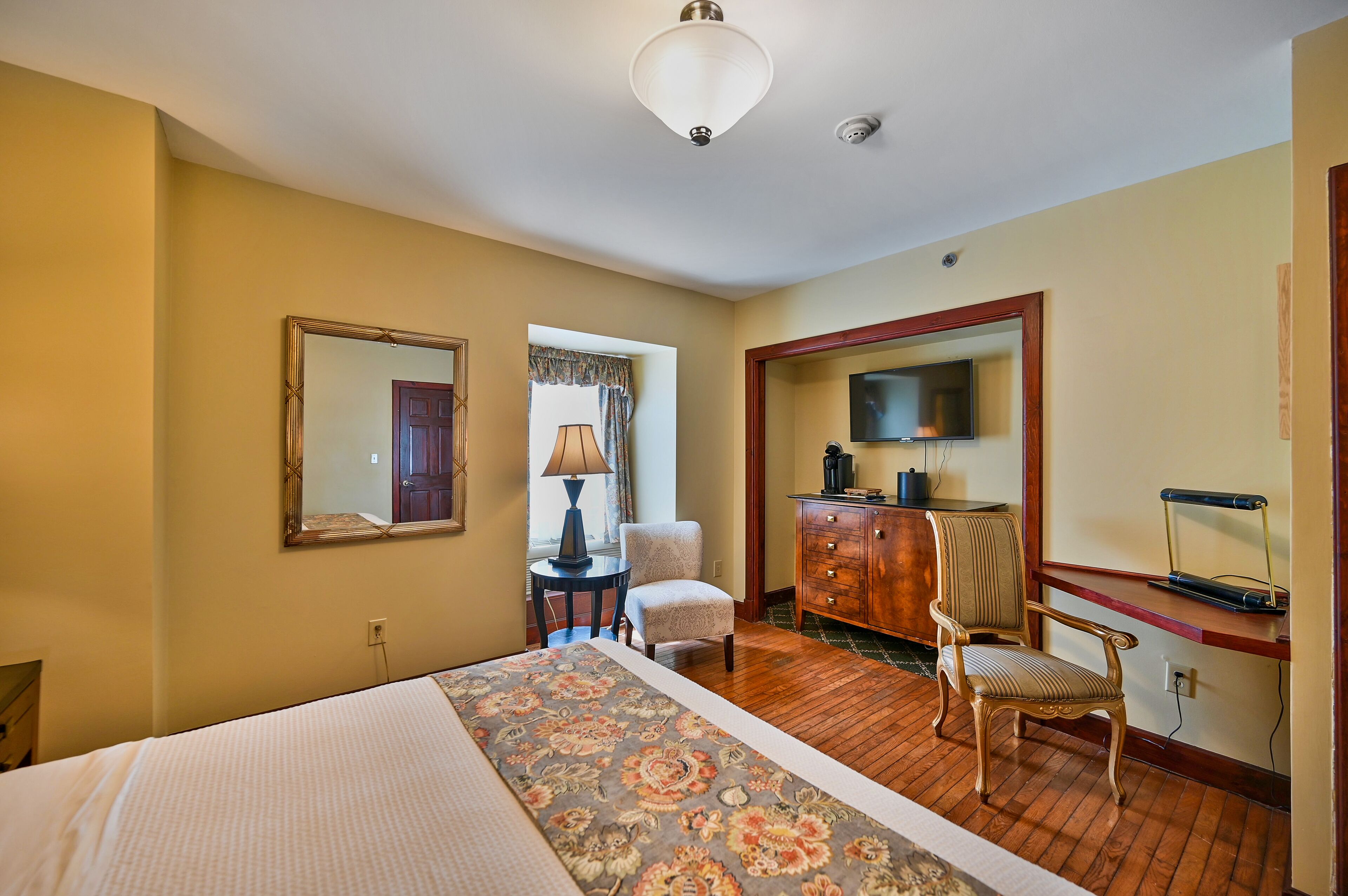 Una habitación de hotel con cama, escritorio con lámpara, silla, aparador y televisor.