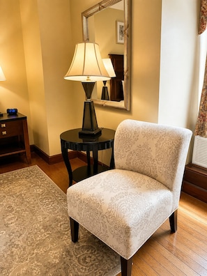 A hotel room with a patterned armchair, a round table with a lamp, a small bedside table with a clock, a mirror, and a patterned rug.