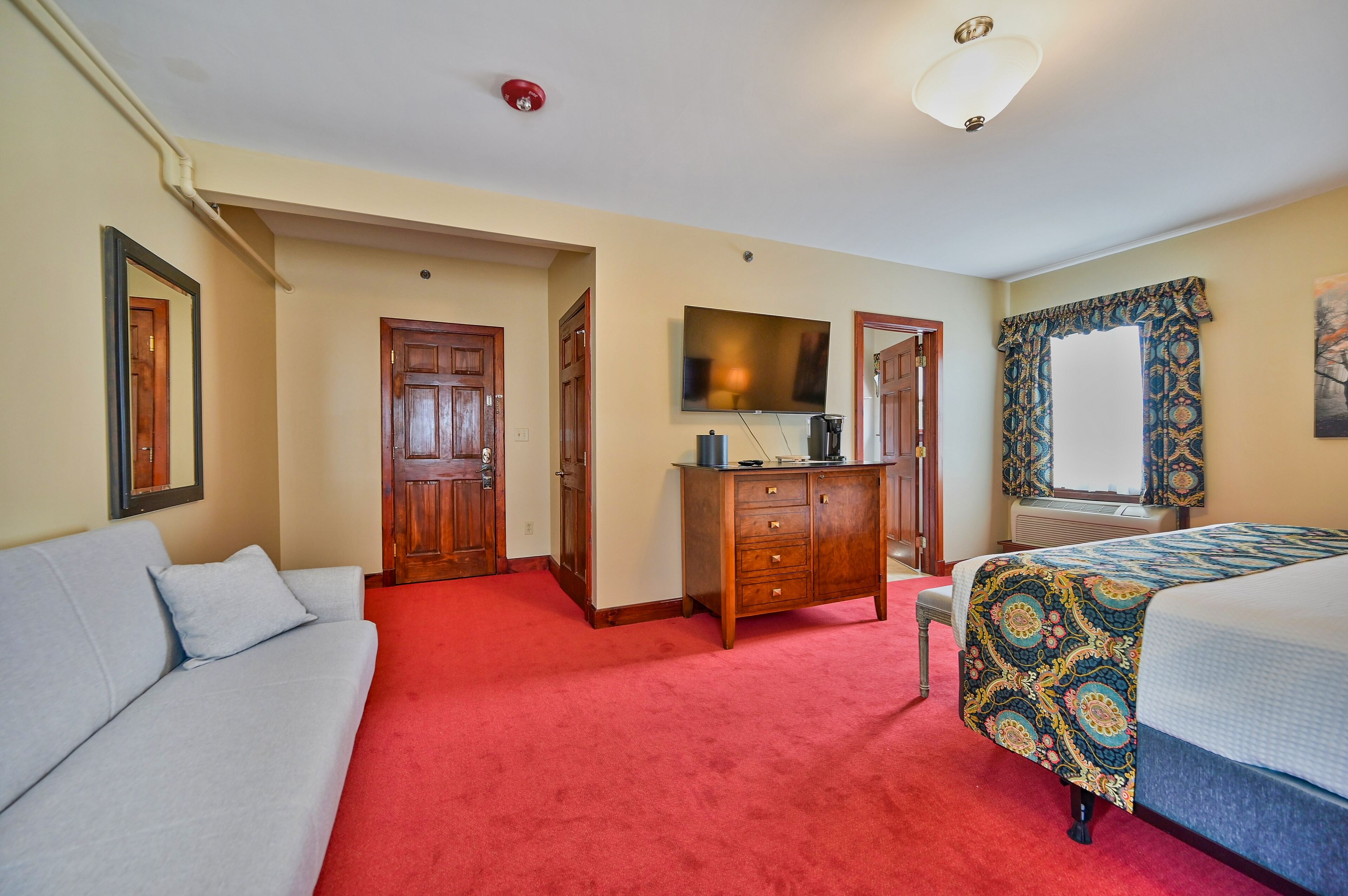 Habitación de hotel con alfombra roja, un sofá, una cama con colcha estampada, un aparador de madera, un televisor de pantalla plana y una ventana con cortinas decorativas.