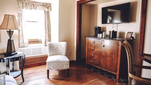 Una habitación con piso de madera, un sillón blanco, un armario de madera, un televisor montado en la pared y una ventana con cortinas.