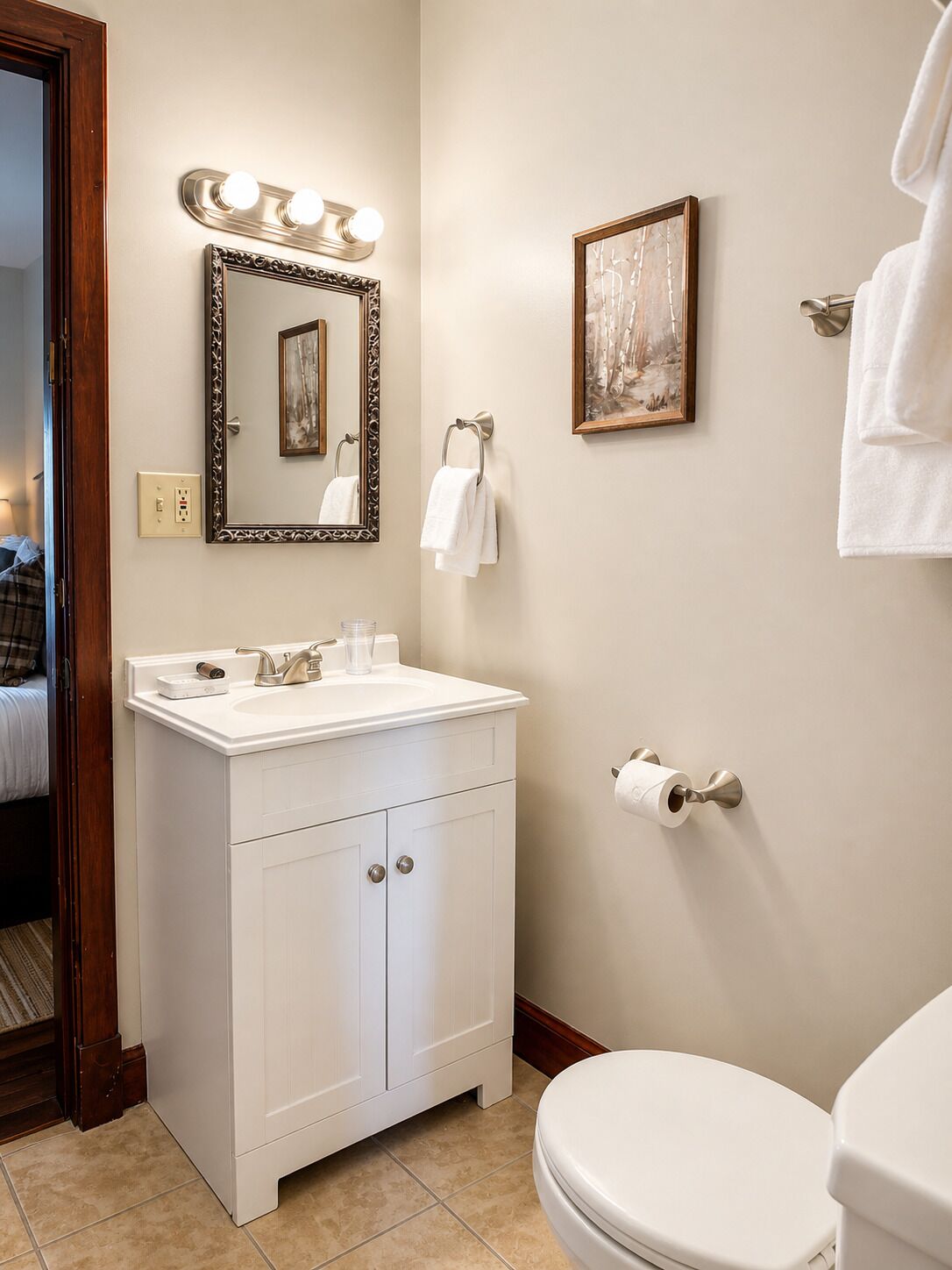A bathroom with a white vanity, a mirror with lights, a framed picture, and a toilet.