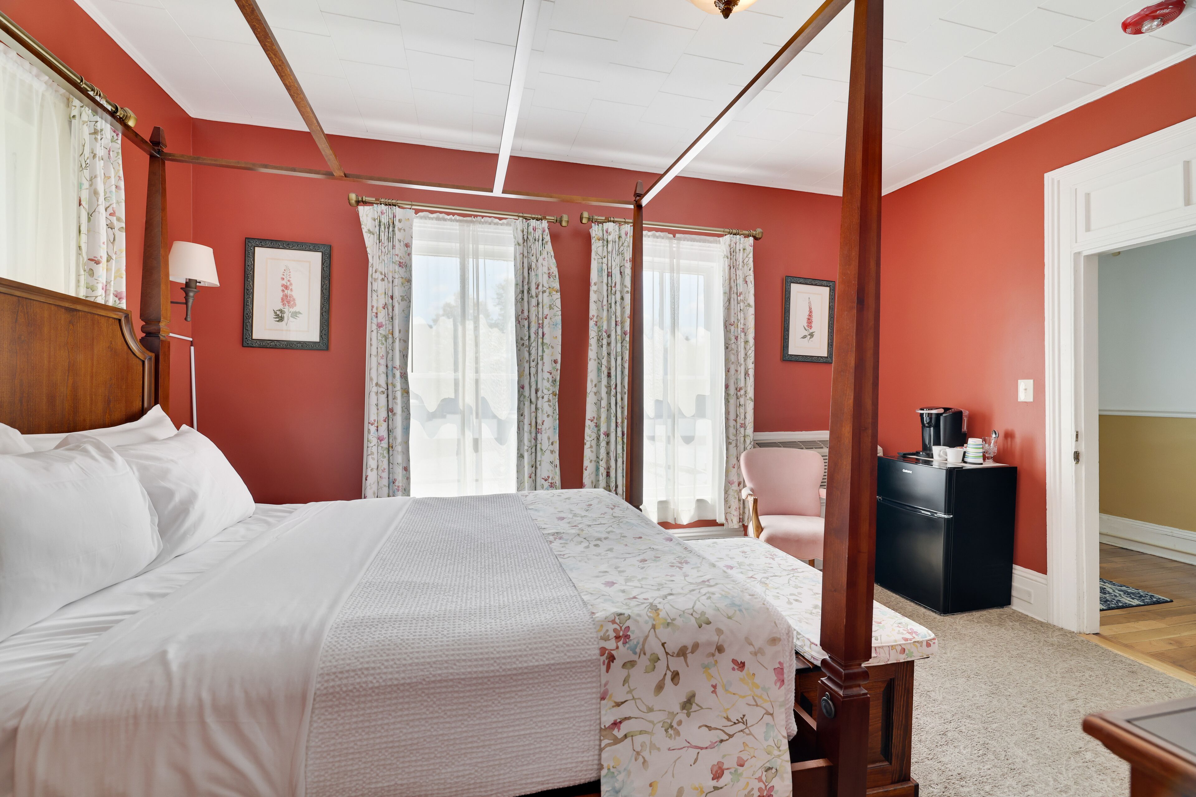 A bedroom with a four-poster bed, floral curtains, a small table with a lamp, and a refrigerator in the corner.