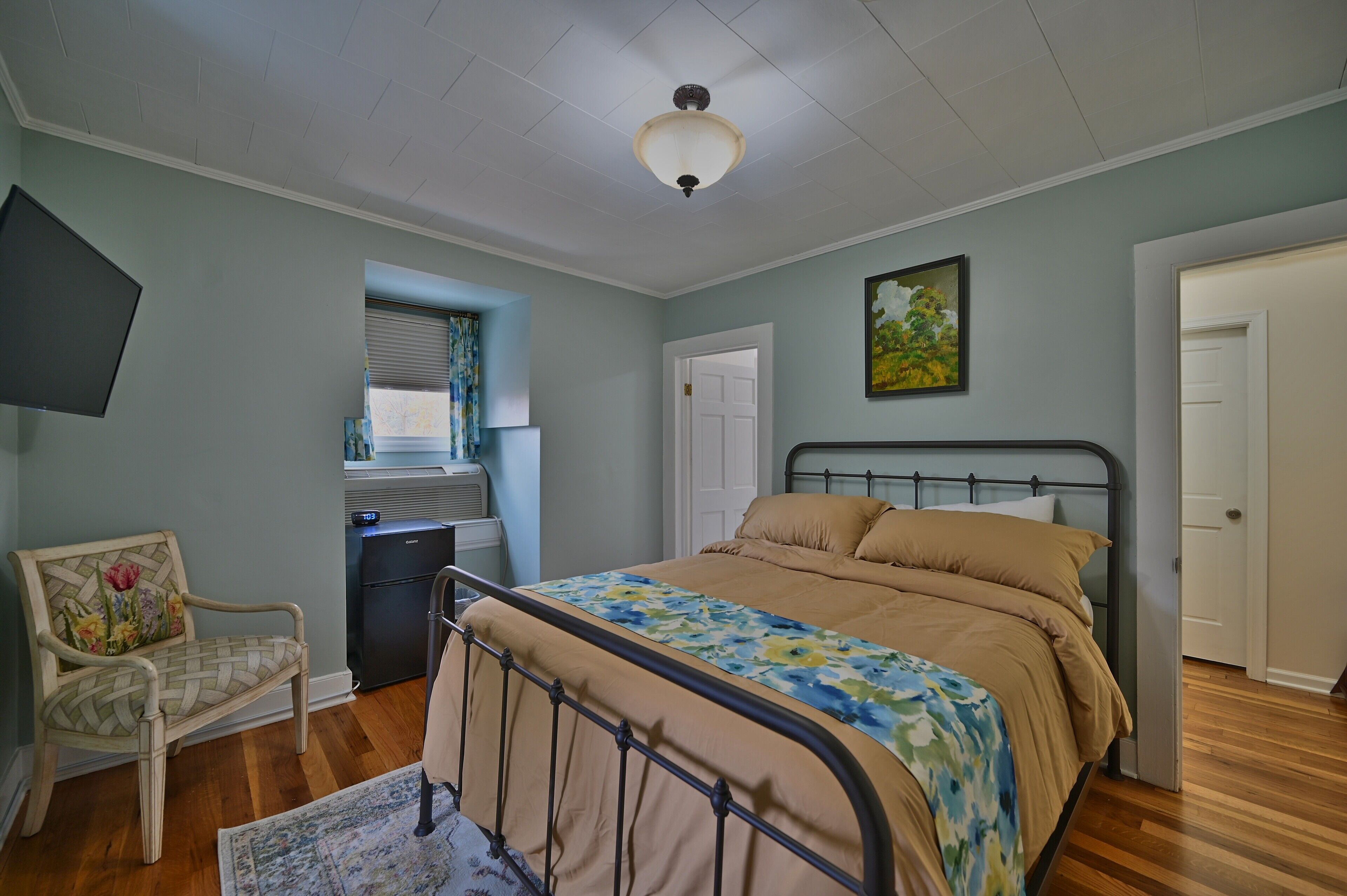 A bedroom with a bed, a chair, a desk, a window with blinds, and a door.