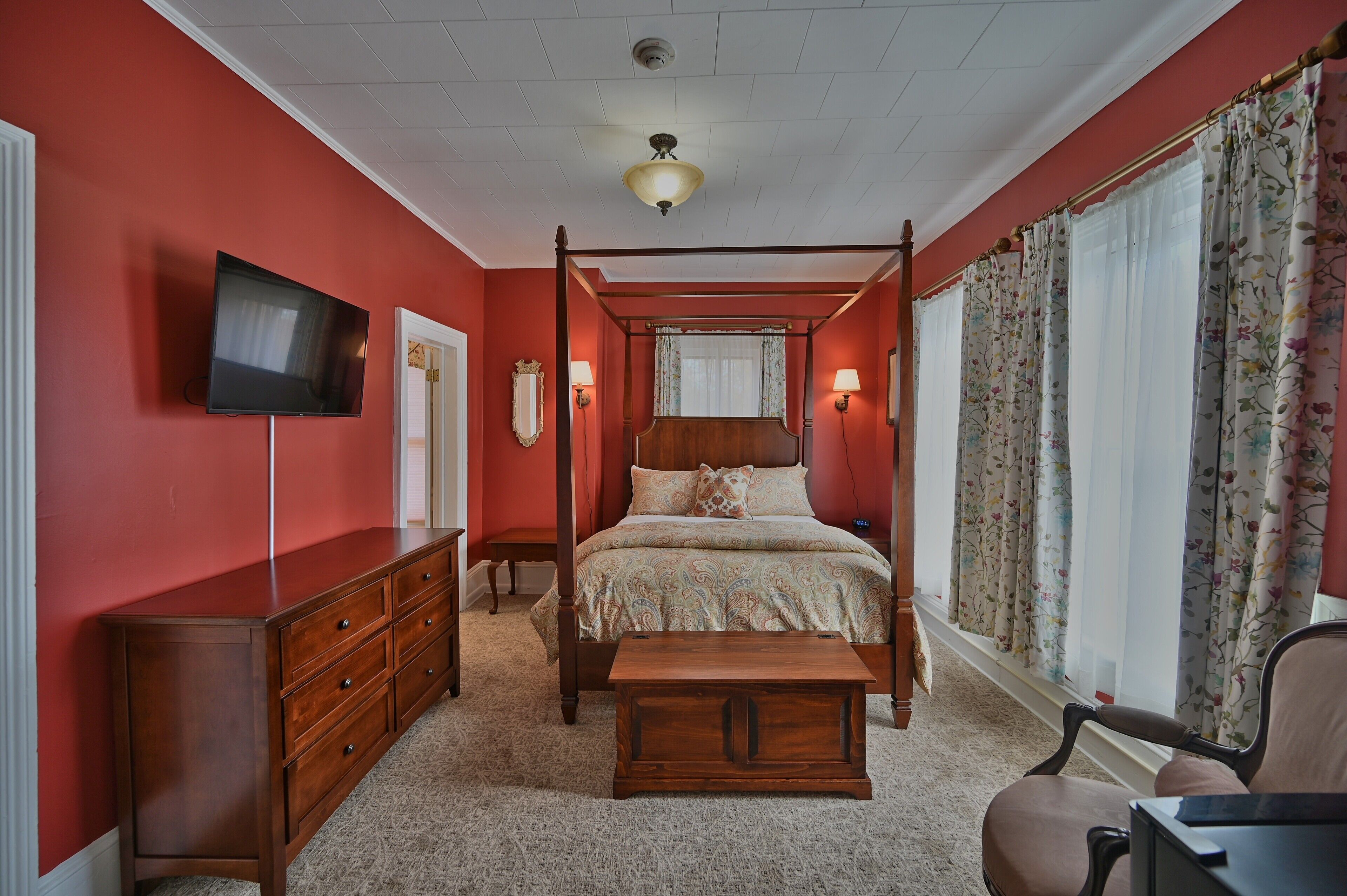 Un lit à baldaquin avec une tête de lit en bois, un téléviseur à écran plat fixé au mur, une commode en bois et une chaise.