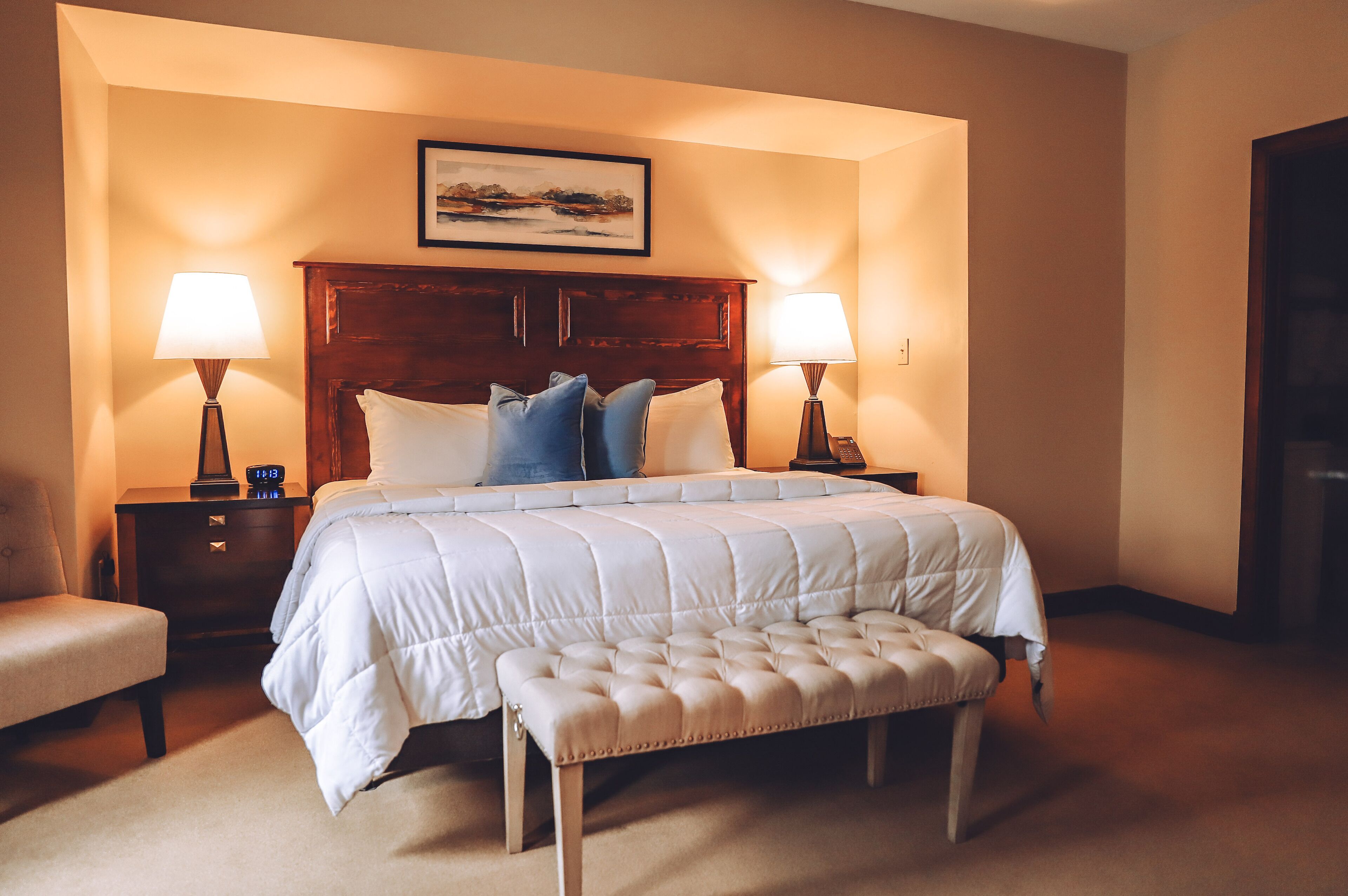A hotel room with a large bed, two bedside tables with lamps, a headboard, a bench, and a framed picture above the bed.