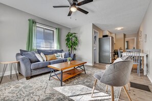 Un salon moderne avec un canapé gris, une table basse en bois, un fauteuil gris et un ventilateur de plafond.