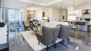 Un salon moderne avec une table basse en verre, deux fauteuils gris, un canapé blanc et un coin repas comprenant une table et des chaises en verre.