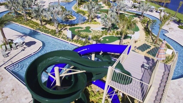 Un parc aquatique doté d’un grand toboggan vert, de palmiers et d’un espace piscine.