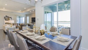 Un espace repas avec une longue table dressée pour six personnes, un lustre et une vue sur la mer grâce à de grandes fenêtres.