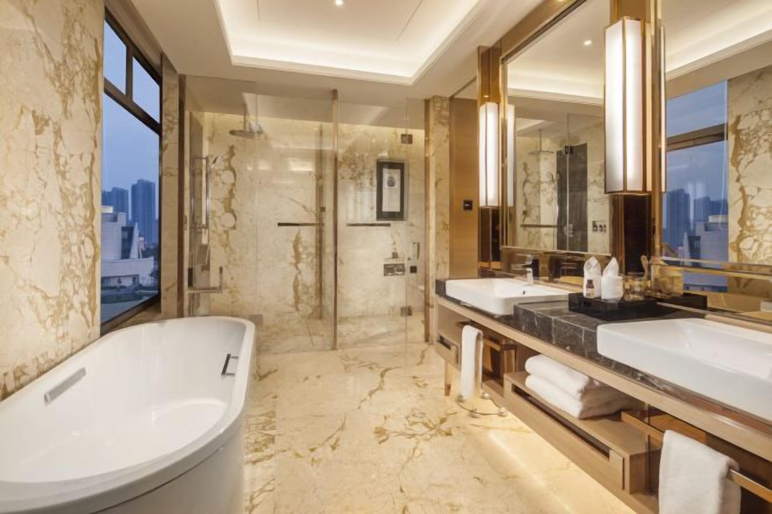 A modern bathroom with a large bathtub, two sinks, a glass shower enclosure, and a city view.