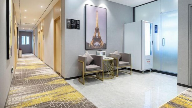 Un couloir d’hôtel avec un tapis à motifs, une grande affiche de la Tour Eiffel, un mini-bar et une porte dotée d’un écran numérique.