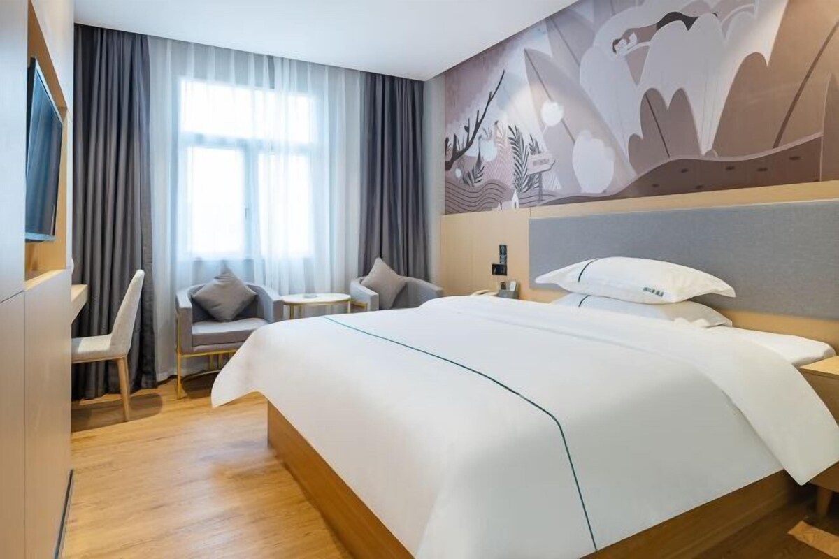 Une chambre d’hôtel moderne avec un grand lit, un bureau et une chaise. On y trouve une fenêtre avec des rideaux, un téléviseur fixé au mur et une fresque décorative.