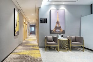 Un couloir d’hôtel avec un tapis à motifs, un coin salon avec deux fauteuils et une table, et une grande image encadrée de la Tour Eiffel accrochée au mur.