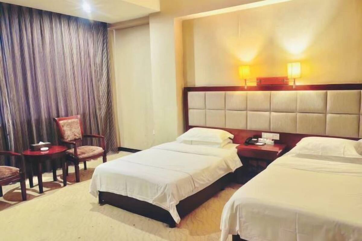 A hotel room with two beds, a chair, a small table, and a curtain.