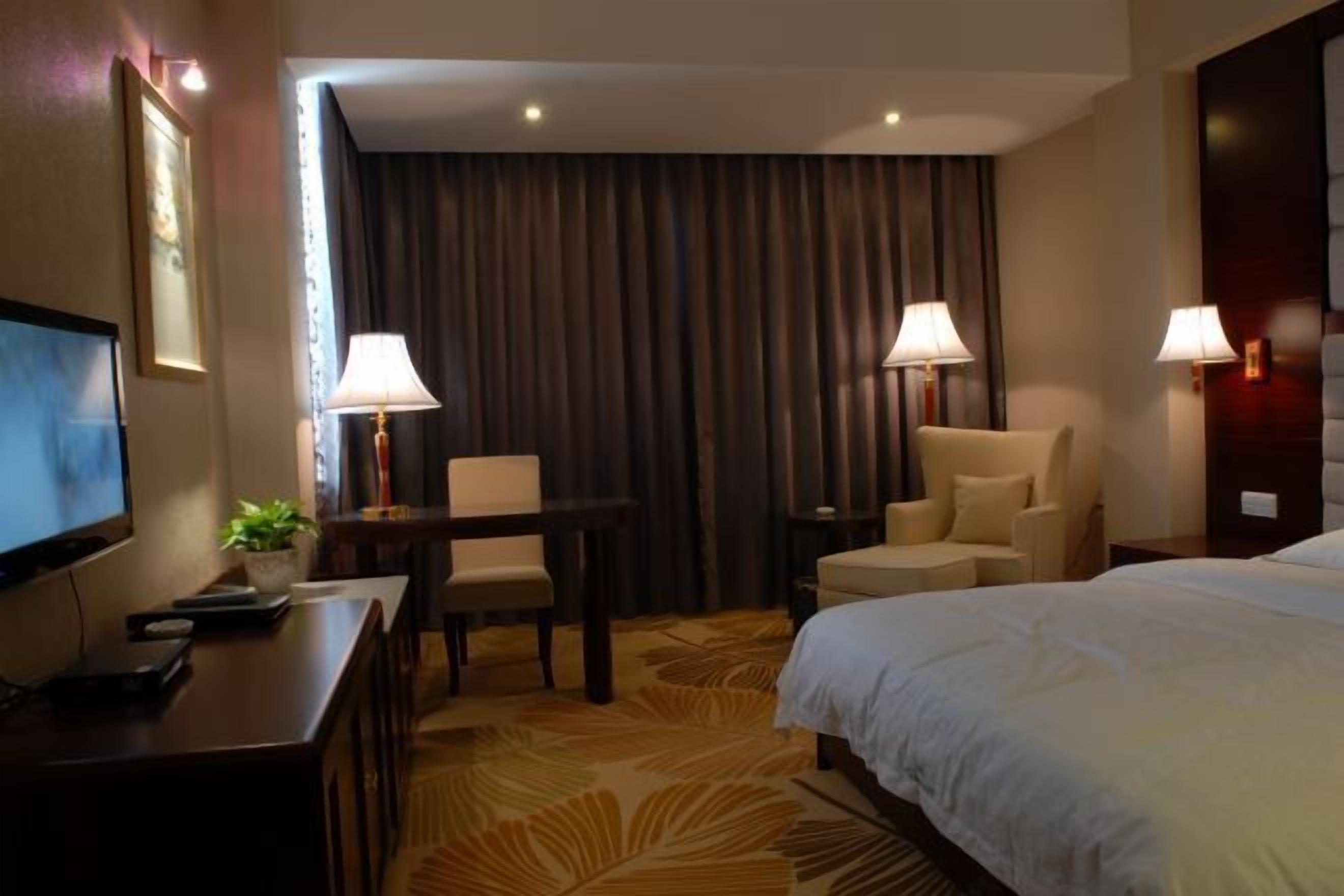 Habitación de hotel con cama, dos sillones, escritorio con lámpara, televisor y un gran ventanal con cortinas.
