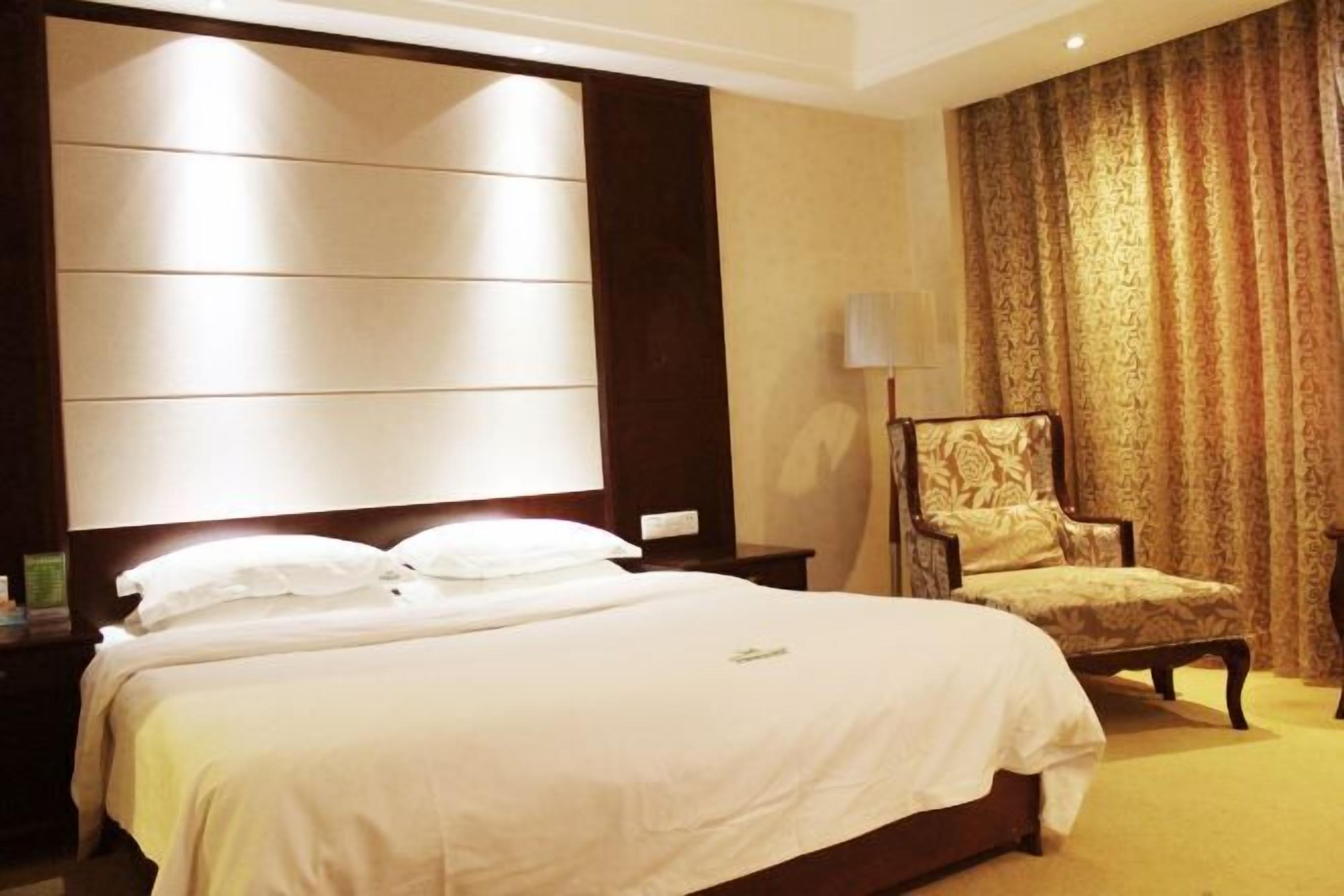 Habitació d'hotel amb un llit gran, una petita taula de nit, una cadira i una finestra gran amb cortines.