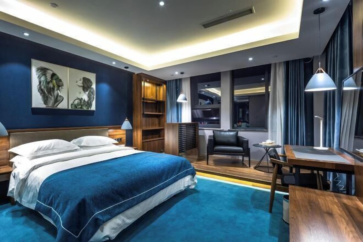 A modern hotel room with a large bed, a desk, a chair, a bookshelf, and a window with curtains.
