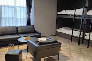 Una habitación de hotel moderna con sofá, una mesita redonda pequeña, una silla, una cama litera y una ventana con cortinas.