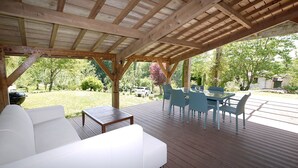 A covered outdoor patio with a white sofa and blue chairs, a wooden deck, and a view of a garden and trees. - Quiet house with large garden and pool, in the Entre-deux-Mers (Saint-Vincent-de-Pertignas)