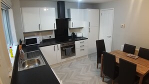 A compact kitchen with white cabinets, a black range hood, a microwave, and a toaster. There's a sink with a faucet, a wooden dining table with chairs, and a herringbone-patterned floor. - Berlin Town house Belfast (Belfast)