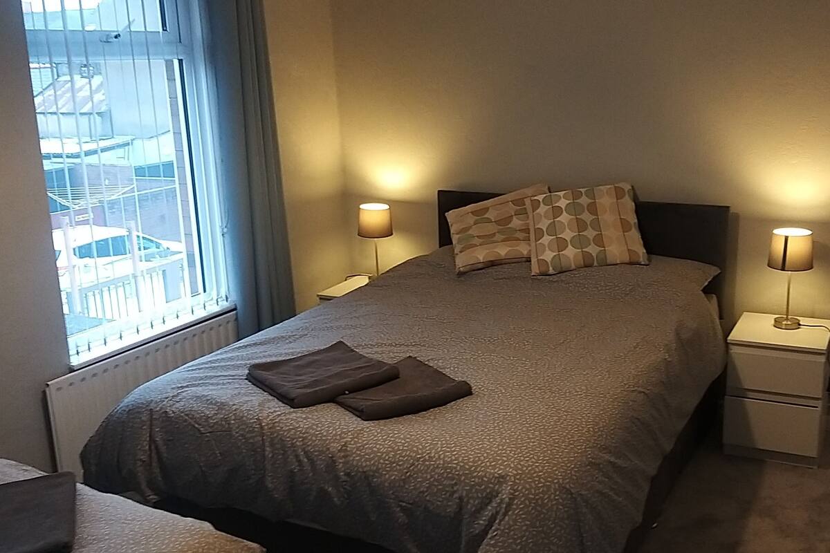 A neatly made bed with a grey blanket and two pillows, two bedside lamps, and a window with a view of buildings.
