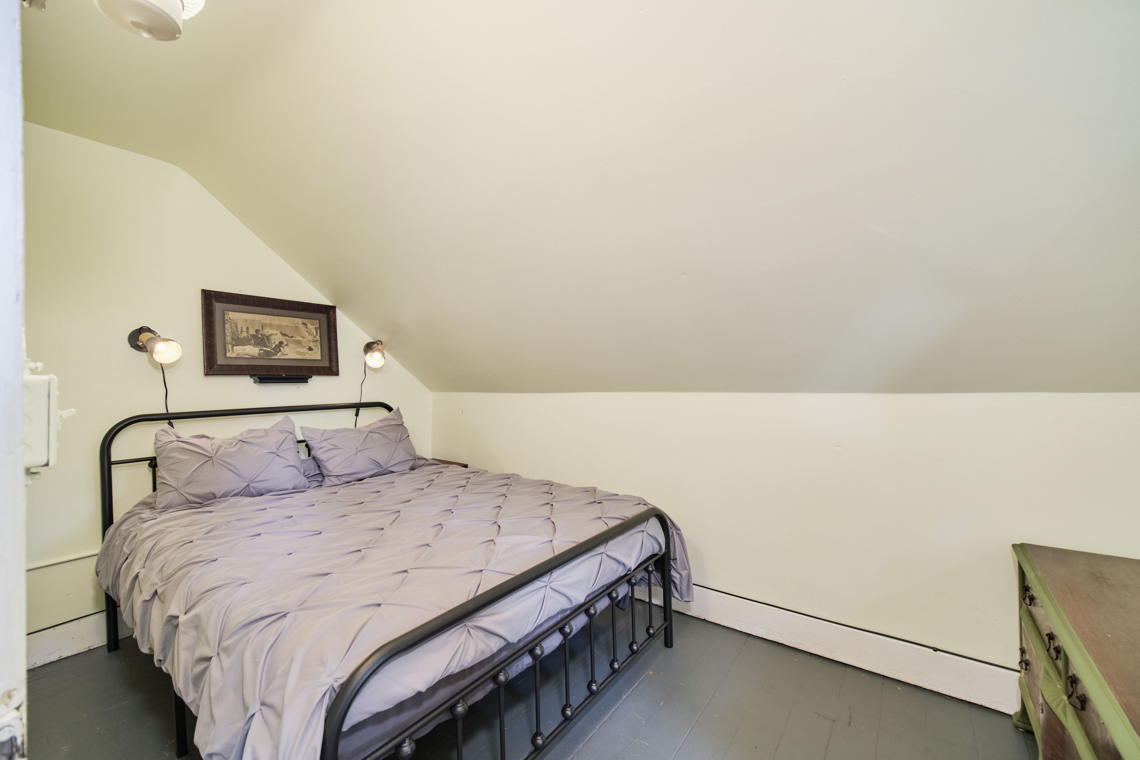 A single bed with a metal frame, a headboard, and a grey bedspread. There is a framed picture on the wall above the bed.