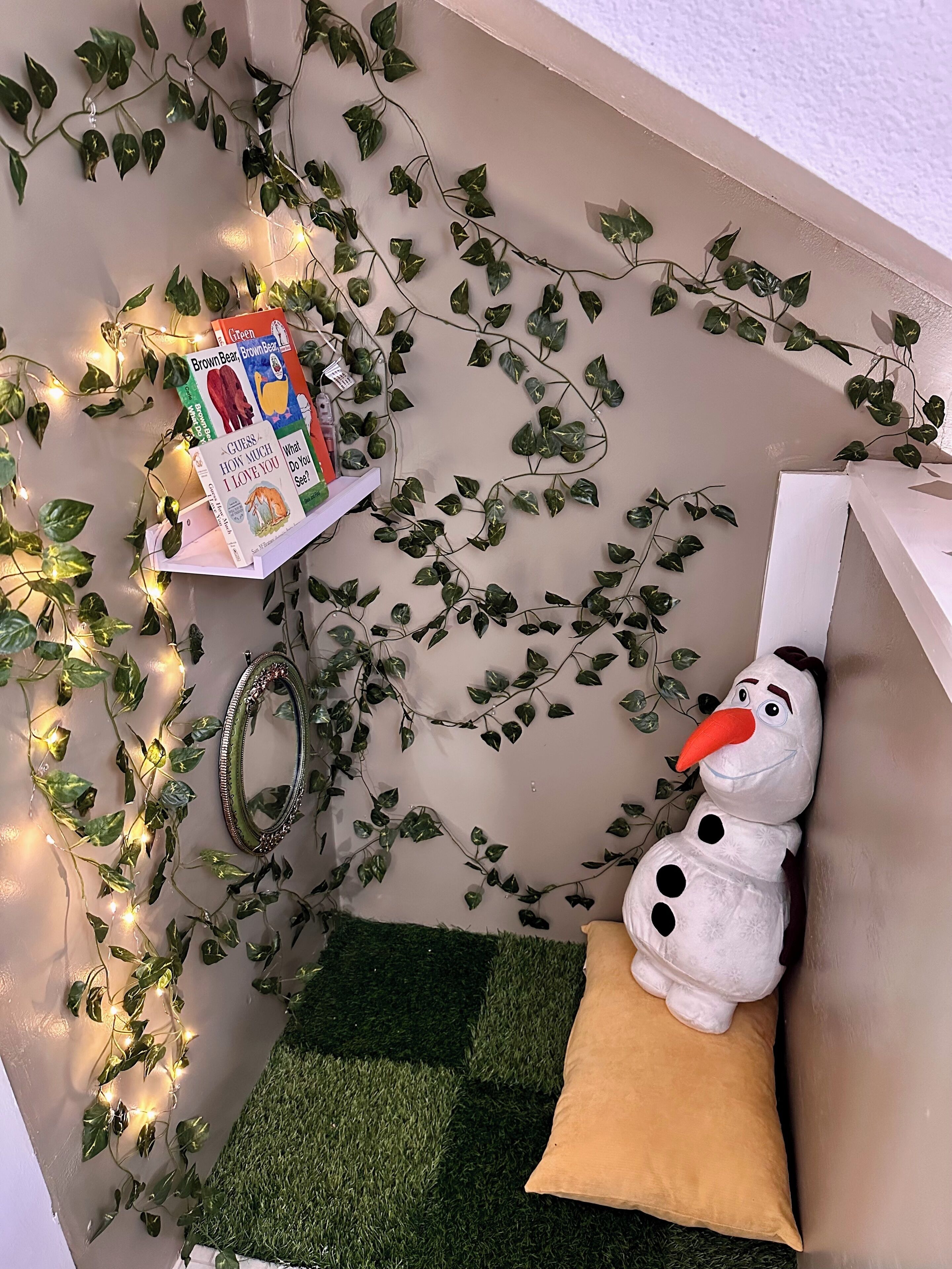 Een hoek van een kamer met een decoratieve plant, een plank met boeken, een spiegel en een zacht knuffeldiertje.