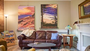 Una sala de estar acogedora con un sofá de cuero, una mesa de centro y una chimenea. Hay dos cuadros grandes en la pared, uno que representa un atardecer sobre un lago y el otro muestra un paisaje de bosque. Se ve una estantería llena de libros en una esquina.