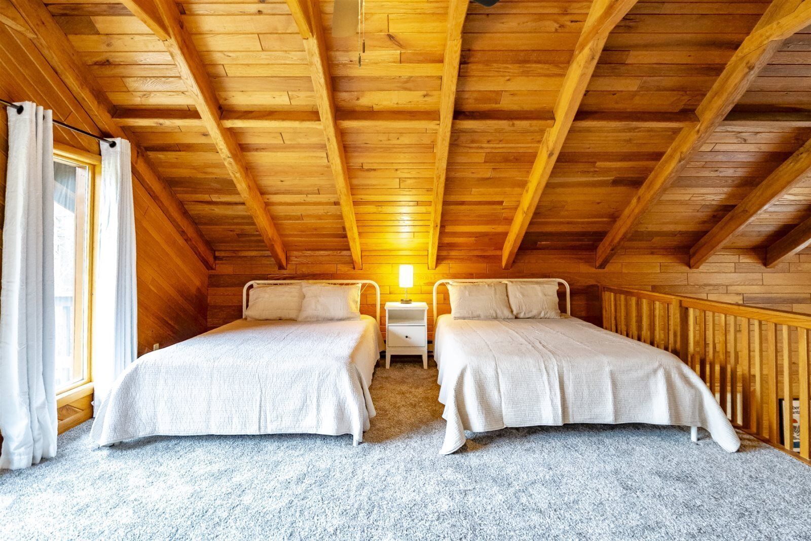 A cozy attic bedroom with two beds, wooden beams, a window with curtains, and a nightstand with a lamp.