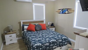 A bedroom with a bed, nightstands, a lamp, a wall clock, and a window with blinds. - BROWNY'S R & R - Beach Cottage (South West Rocks)
