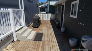 A wooden deck with steps leading to a covered area, a white fence, and a small outdoor seating area with chairs and a table. - BROWNY'S R & R - Beach Cottage (South West Rocks)