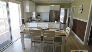A modern kitchen with white cabinets, a stainless steel refrigerator, and a dining area with a wooden table and two chairs. - BROWNY'S R & R - Beach Cottage (South West Rocks)