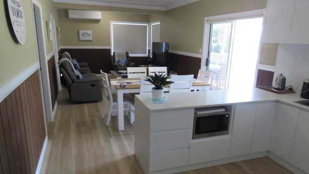 A modern kitchen with a central island, white cabinetry, and a dining area with a sofa and chairs. - BROWNY'S R & R - Beach Cottage (South West Rocks)