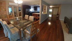 Zona menjador àmplia amb taula de vidre i cadires de fusta, cuina al fons i un passadís que condueix a una altra habitació.